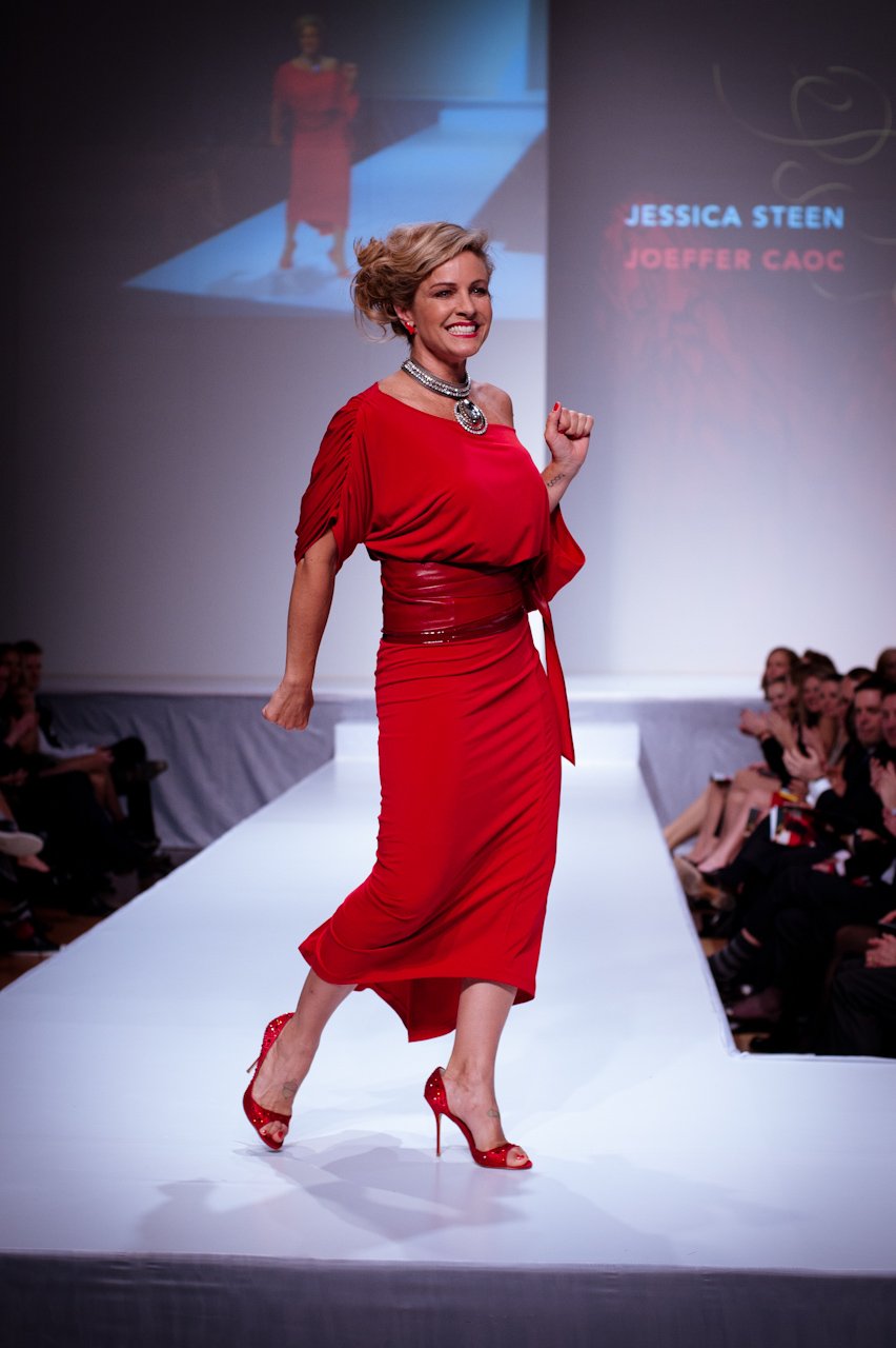 Woman walking a runway in a draped wine-red midi dress with a wide belt, layered statement necklace and red strappy high heels