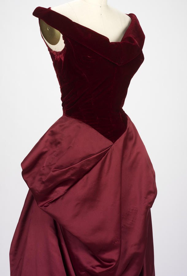 Wine-red velvet and satin evening gown by Charles James, 1955, displayed on a mannequin: fitted velvet bodice with draped off-shoulder neckline and full satin skirt.