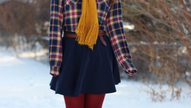 A woman with long brown hair wearing a plaid shirt jacket, a mustard yellow scarf, a dark skirt, red tights and brown boots, standing in the snow