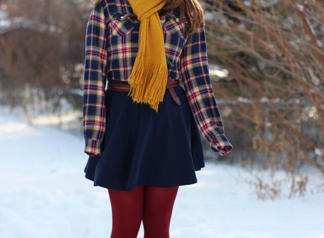 A woman with long brown hair wearing a plaid shirt jacket, a mustard yellow scarf, a dark skirt, red tights and brown boots, standing in the snow