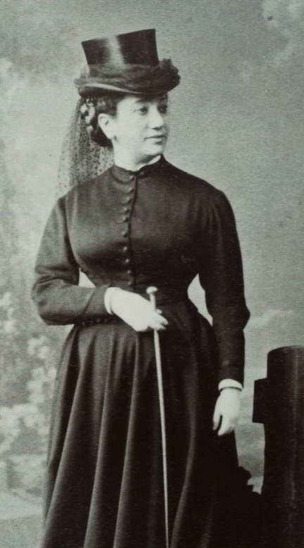 1860s black-and-white portrait of a woman in a dark buttoned riding-habit dress and tall top hat with veil, holding a riding crop