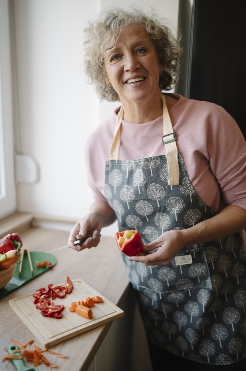 A smiling woman with curly grey hair wearing a pink top and a grey tree-print apron, chopping red peppers on a wooden cutting board in a bright kitchen