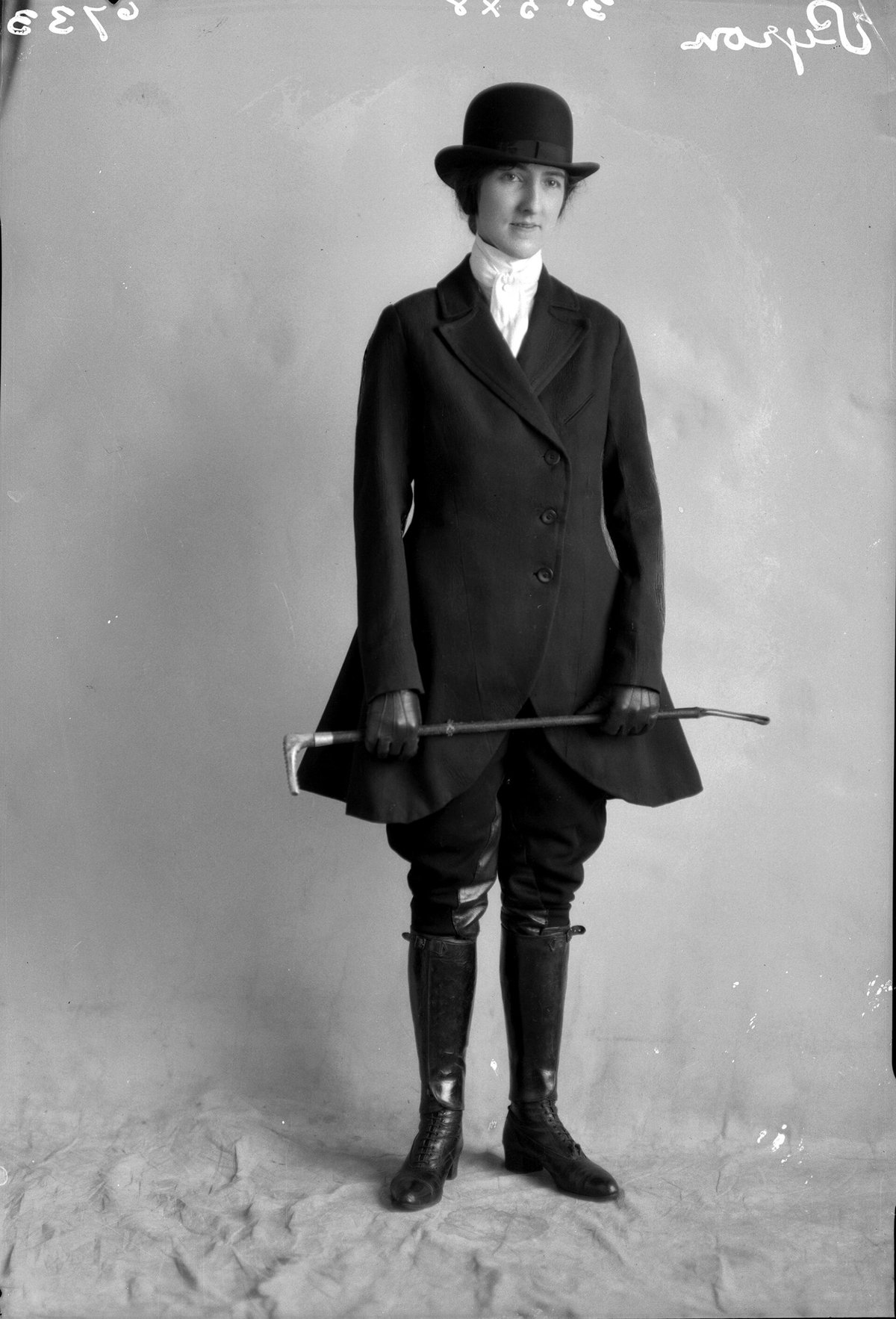 Black-and-white studio portrait of a woman in a full black riding habit with a tailored long coat, breeches, tall lace-up riding boots, a white stock and a bowler hat, holding a riding crop
