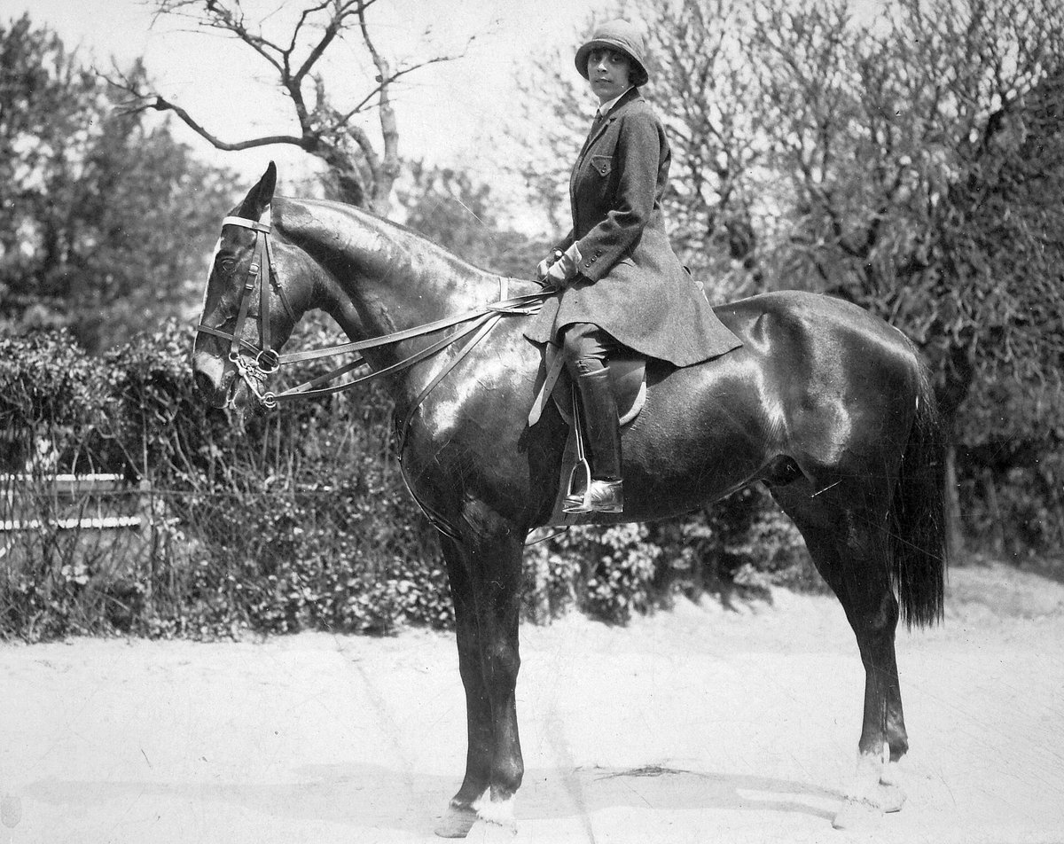 Black-and-white photograph of a woman on horseback among bare trees, wearing a riding coat, tall boots and a bowler hat