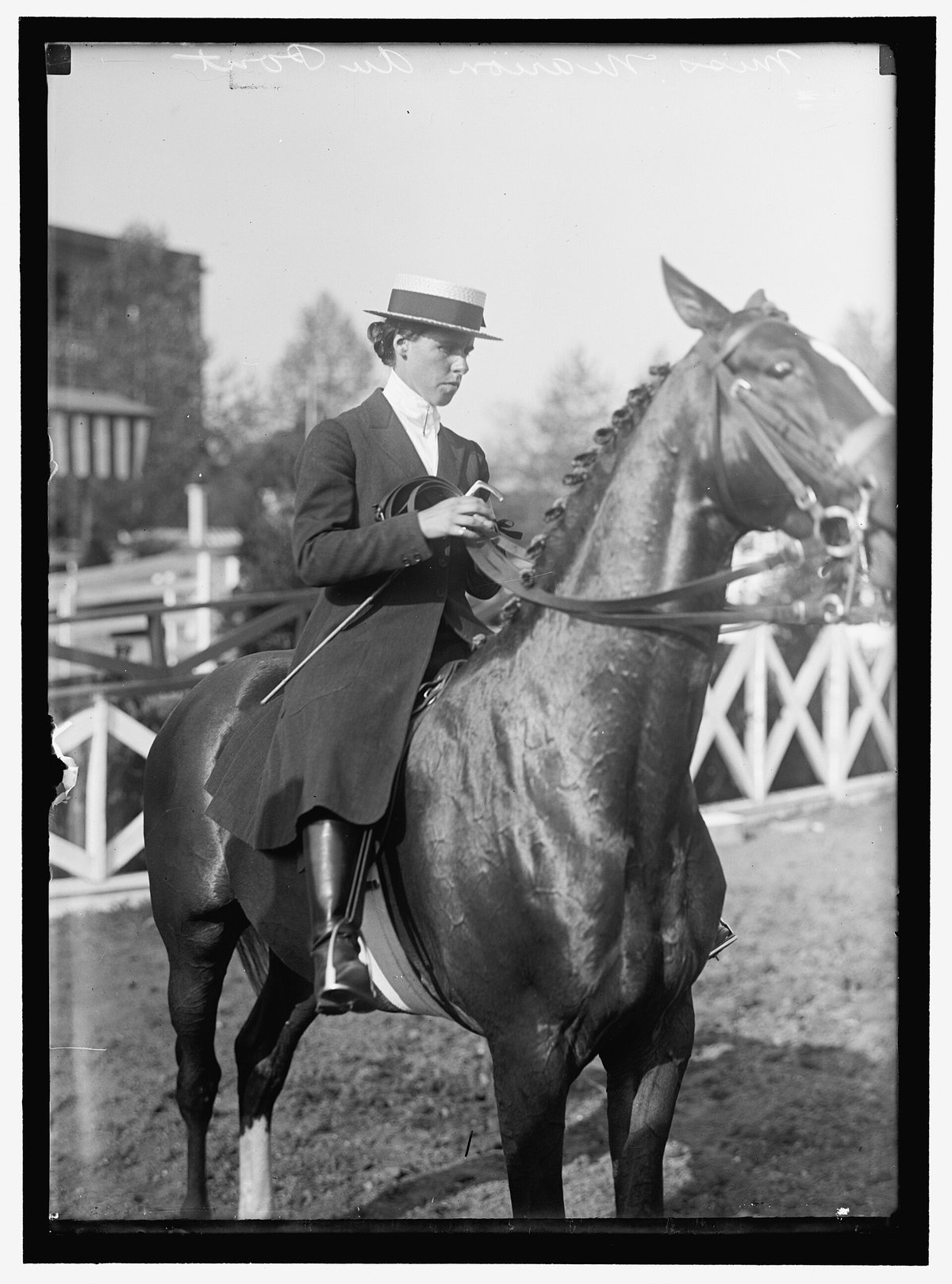 Black-and-white photograph of a woman mounted on a horse at a horse show, wearing a dark tailored riding coat, white stock, tall black boots and a straw boater hat