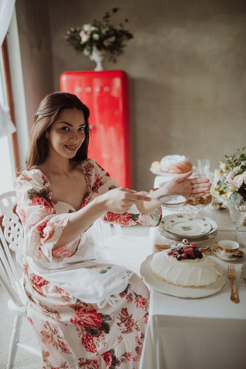 A young woman in a floral dress and white lace-trimmed apron, dusting powdered sugar over a berry-topped dessert at an elegantly set dining table