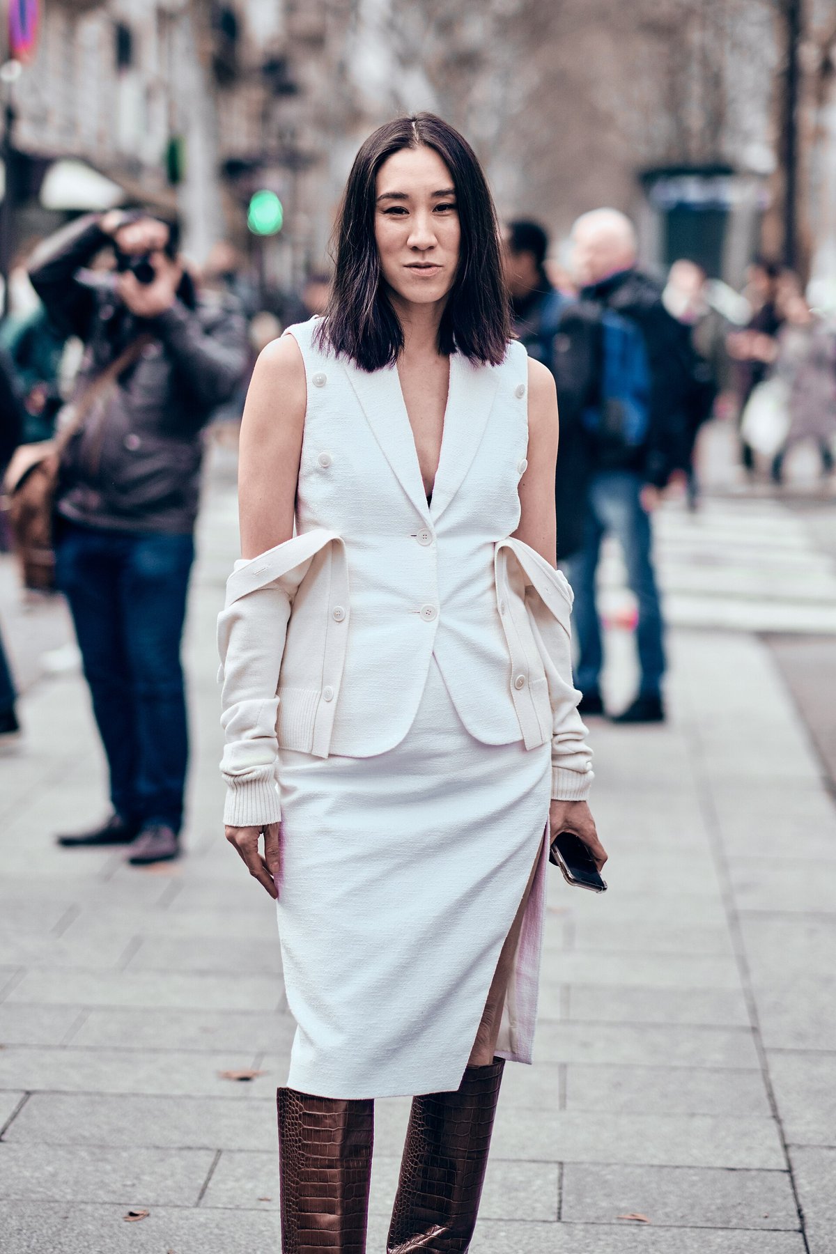 Color street-style photograph of a woman in a cream sleeveless blazer and white slit skirt wearing tall brown croc-embossed knee-high boots