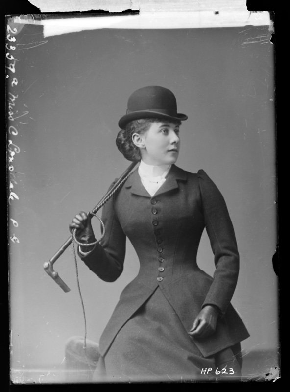 Black-and-white Victorian studio portrait of a woman in a buttoned riding-habit jacket and bowler hat, holding a whip and riding crop
