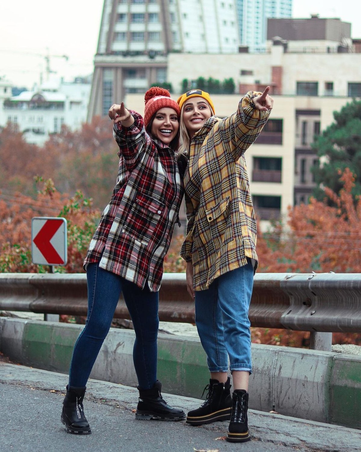Two women outdoors in autumn, one wearing a yellow plaid shacket with a yellow beanie and the other a red plaid shacket, both in jeans and black boots