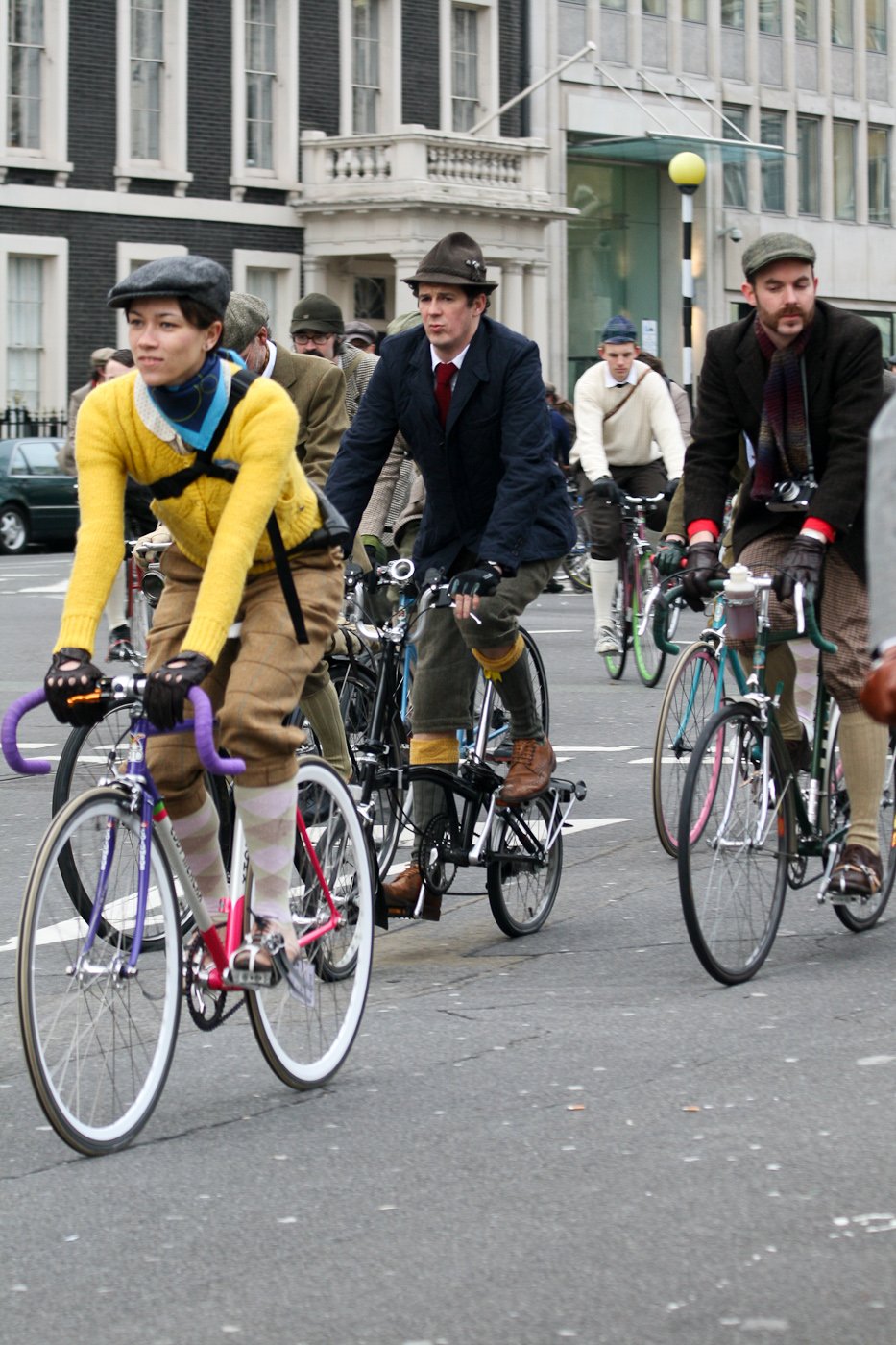 Woman riding a bicycle in a mustard-yellow sweater, brown tweed knickerbockers, patterned socks and a tweed flat cap.